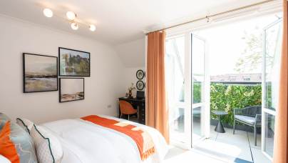 A bright bedroom with a bed, desk, and wall art, featuring open glass doors leading to a balcony with greenery and outdoor seating.