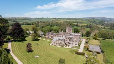 Aerial view of a large stone mansion surrounded by expansive green lawns, trees, and countryside under a clear blue sky.