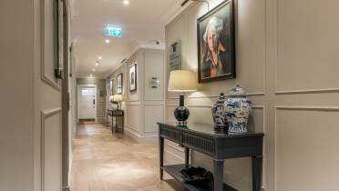 A hallway with wooden floors, neutral walls, framed portraits, a ceramic vase, a table lamp, and a console table under warm lighting.