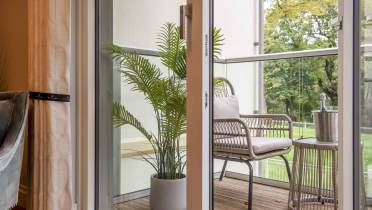 A balcony with wicker seating and a potted plant is seen through an open glass door. A small table holds a champagne bottle in an ice bucket. Trees are visible in the background.