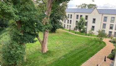 View of a grassy courtyard with large trees, a paved walkway, and a modern white three-story building in the background under a cloudy sky.