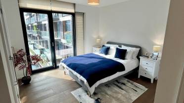 Modern bedroom with a double bed, blue and white bedding, two bedside tables, a rug, a potted plant, and large window overlooking a building.