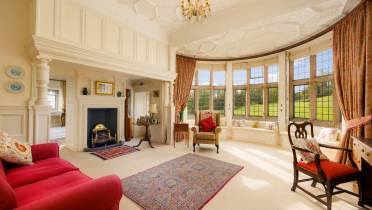 Elegant living room with large bay windows, red sofas, an ornate rug, and a fireplace. A chandelier hangs from a detailed ceiling, and the room overlooks a garden.