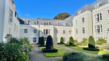 A courtyard with trimmed hedges and a walkway is surrounded by multi-story white buildings under a clear blue sky.