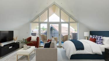 A spacious bedroom with a vaulted ceiling features a bed, a TV, a red couch, a beige loveseat, and a small balcony with outdoor chairs, all under large triangular windows.