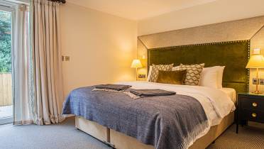 A neatly arranged bedroom with a large bed. There are lamps on nightstands, a large window with curtains, and a gray carpet covers the floor.