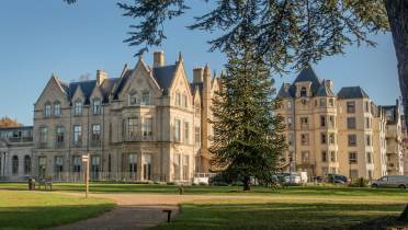 A large historic building with ornate architecture is surrounded by a grassy area, trees, and a clear blue sky.