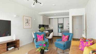 Modern living room with two blue armchairs, a yellow sofa, and a wooden coffee table. Bright pillows and plants add color. A kitchen is visible in the background, and a sleek light fixture hangs overhead.