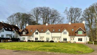 Row of white houses with brown roofs, set in front of leafless trees, facing a circular lawn area.