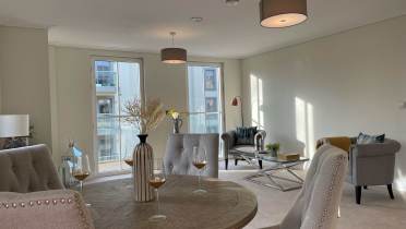 Bright living and dining area with modern decor, featuring a round table, gray upholstered chairs, a sofa, armchairs, and large windows with views of adjacent buildings.