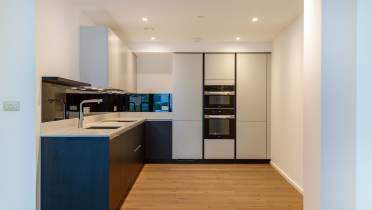 Modern kitchen with wooden floors, white walls, and recessed lighting. Features a sleek black counter, built-in double oven, and minimalist cabinets.