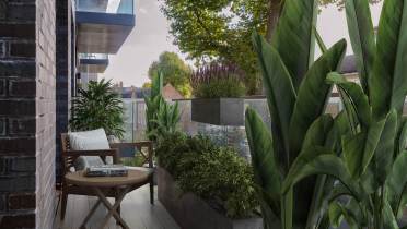 A modern balcony with lush plants, a wooden chair, and a small round table holding a book. A clear glass railing overlooks suburban houses and green trees.