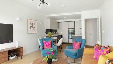 Modern living room with blue and yellow chairs, a wooden coffee table, and a wall-mounted TV. A kitchen is visible in the background, and bright cushions add color to the space.