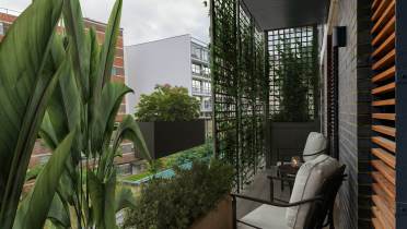 A small urban balcony with a chair, side table, and plants. Vertical trellis with greenery partially obstructs the view of neighboring buildings.