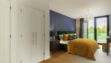 Modern bedroom with a large bed, mustard duvet, green upholstered headboard, and wooden flooring. There are white closets, a navy accent wall, and a window with a view of greenery.