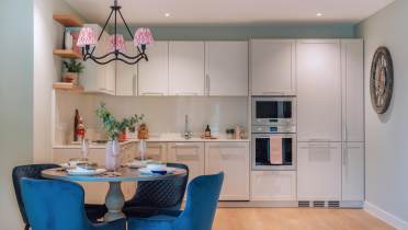 Modern kitchen featuring white cabinets, built-in ovens, a round dining table with blue chairs, and a black chandelier with pink shades. A large wall clock is visible.