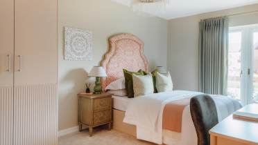 A neatly arranged bedroom featuring a bed with orange patterned headboard, green and white pillows, bedside table with lamps, and a window with drapes. A decorative wall piece hangs above.