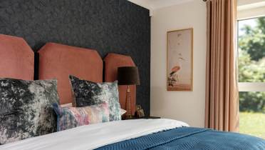 Bedroom with a bed featuring a blue quilt, patterned pillows, a brown headboard, a black lamp on a side table, dark textured wallpaper, and a framed bird print. Curtains cover a window.