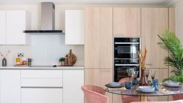 Modern kitchen with white cabinets, light wood paneling, and a stainless steel oven. There is a round dining table with pink chairs and tableware. A large green plant is in the corner.