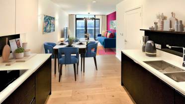Modern open-plan kitchen and living area with wood flooring, black and white cabinetry, a dining table with blue chairs, and a blue sofa. Colorful artwork and decor accent the space.