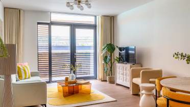 Modern living room with large windows, a light sofa, orange coffee table, yellow rug, and a TV on a white cabinet. A round dining table with yellow chairs is in the corner.