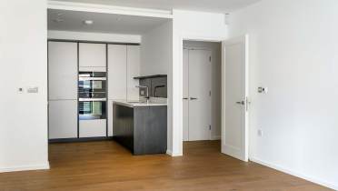 Modern kitchen with built-in appliances, sleek cabinets, and a small peninsula. The kitchen is adjacent to an open area with wooden flooring and a partially open white door.