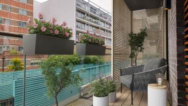 Modern balcony with two planters of pink flowers, a gray cushioned chair, a small round table with a candle, and a potted plant. Wood slat wall and glass railing overlook neighboring buildings.