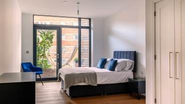 Modern bedroom with a large window, double bed, blue chair, and a hammock visible outside. White walls, wooden floor, and built-in closets complete the minimalist decor.