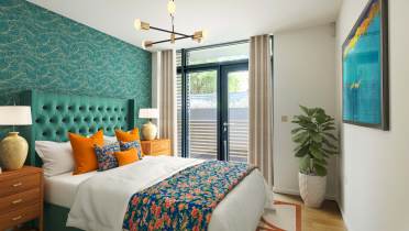 Modern bedroom with a teal accent wall, double bed with colorful linens, two wooden nightstands, a potted plant, abstract art, and a large window with curtains.
