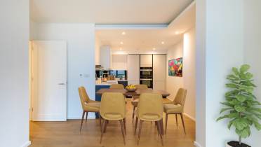 Modern dining area with six mustard-yellow chairs around a wooden table. A kitchen with white cabinets and colorful wall art is visible in the background. A potted plant is on the right.