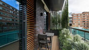 Modern balcony with a wooden table and two chairs, surrounded by greenery. Slatted wood panels and glass railings contrast the brick walls and nearby buildings.