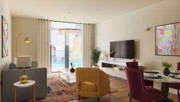 A modern living room features a gray sofa, yellow and pink chairs, a flat-screen TV, and colorful wall art. Large windows overlook a balcony with a pool view.