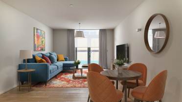 Modern living room with a blue sofa, colorful rug, and orange dining chairs. A round mirror hangs on the wall, and a TV is opposite the sofa. Large window with curtains in the background.