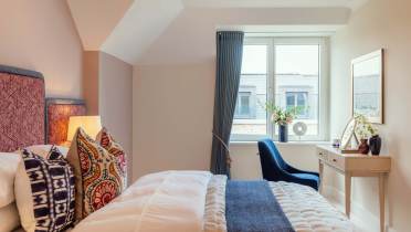 A neatly arranged bedroom with a bed, colorful pillows, a window with curtains, a small desk with flowers, and a blue chair.