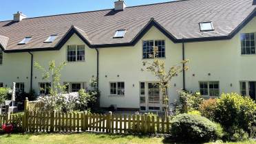 A row of modern, cream-colored townhouses with a brown roof, multiple windows, skylights, and a small fenced garden in the foreground under clear blue sky.