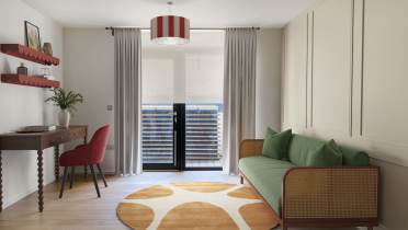 A modern living room with a green sofa, round orange and white rug, a wooden desk with a red chair, and glass doors covered by white curtains leading to a small balcony.