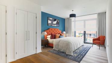 Modern bedroom with an orange upholstered headboard, blue accent wall, white bedding, patterned rug, armchair, two nightstands, and large window with sheer curtains.