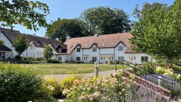 A large white house with a reddish-brown roof is surrounded by gardens, trees, and flowering plants under a clear blue sky.