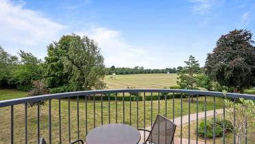 A small round table with two chairs sits on a balcony overlooking a large grassy field and trees under a partly cloudy sky.