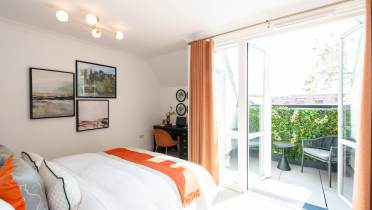 A bright bedroom with a bed, desk, and wall art, featuring open glass doors leading to a balcony with greenery and outdoor seating.