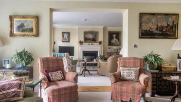 A living room featuring two matching plaid armchairs with personalized pillows, a fireplace in the background, and multiple paintings on the walls.