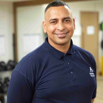 A person in a dark blue polo shirt stands smiling in a room with fitness equipment.