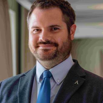 Smiling man in a suit with a blue tie, standing indoors near a window.