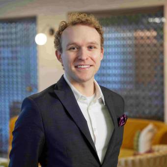 Person with curly hair in a dark suit and white shirt smiles indoors. Background features a modern, softly-lit room with decorative elements.