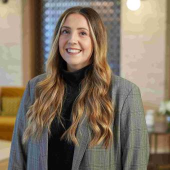 Person with long hair and a plaid blazer smiles at the camera, standing indoors with a patterned backdrop.