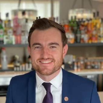 A person in a blue suit and purple tie is smiling at the camera with a background of blurred bottles on shelves.