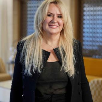 Person with long blonde hair in black clothing stands indoors, smiling.