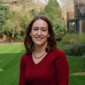 A woman with brown hair wearing a red sweater smiles while standing in a garden with grass and trees in the background.