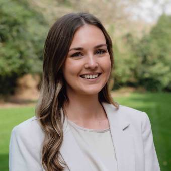 A woman with long brown hair, smiling, wearing a white blazer and shirt. She stands outdoors with a blurred green background.