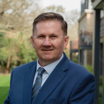 Man in a blue suit and striped tie stands outdoors with trees and buildings in the background.